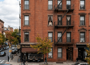 Warmly lit apartment window with plants and a seated person above a city street