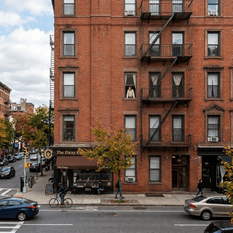 Warmly lit apartment window with plants and a seated person above a city street